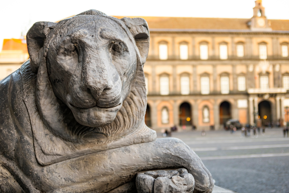 Estatua de león en la Plaza del Plebiscito con el Palacio Real de Nápoles a su espalda